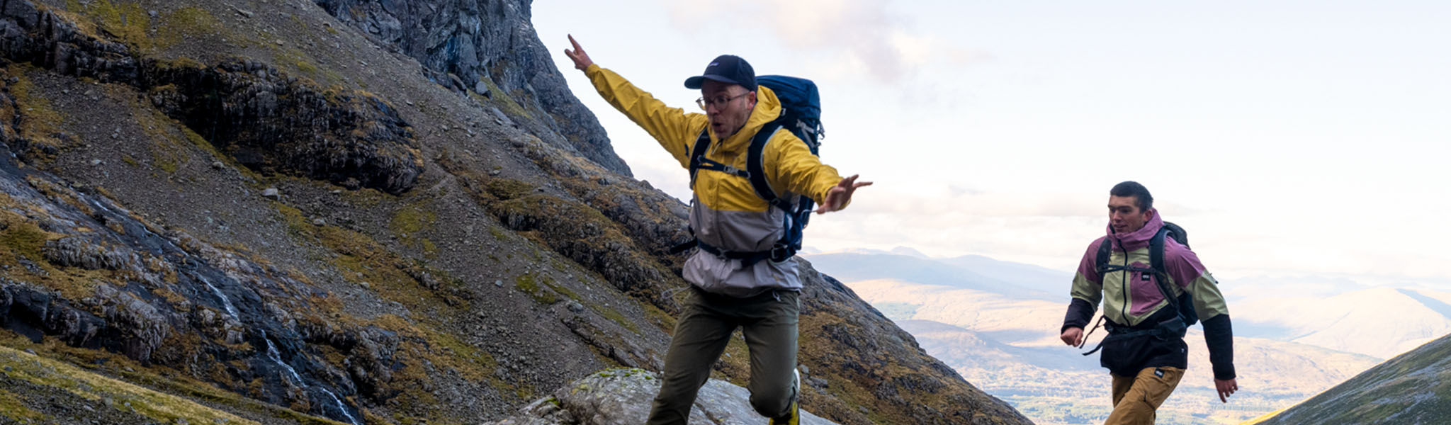 Two men are alking fast on big rocks, wearing walking gear and rucksacks.