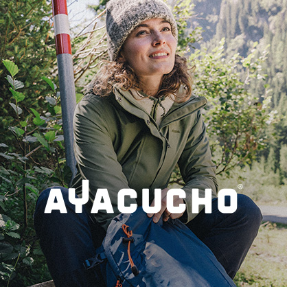 Shop Ayacucho collection. Woman sitting in nature, wearing green jacket and blue troursers