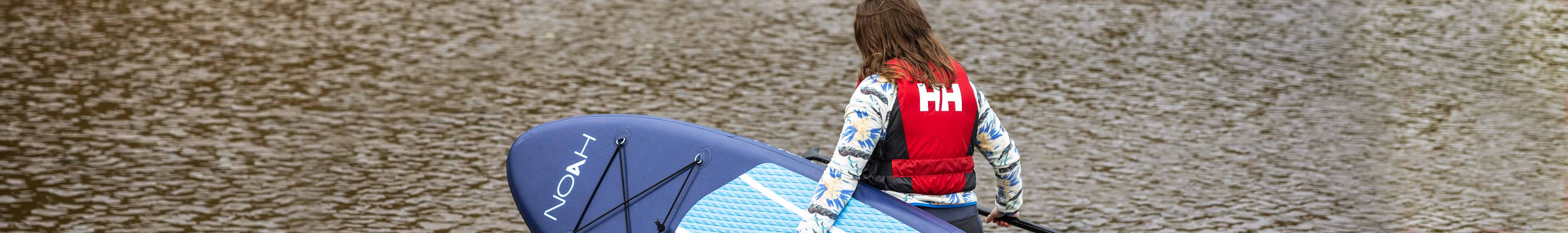 Watersports Spring Summer 23 collection. A woman carrying blue paddleboard on a beach