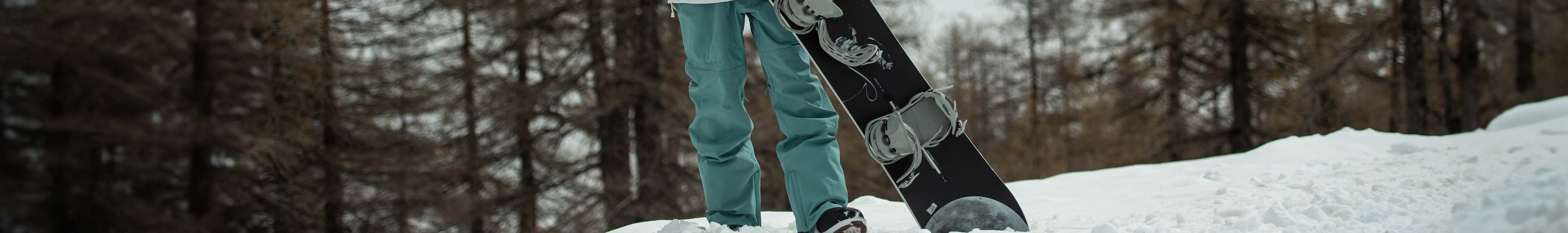 Shop men's legwear. A man wearing snow pants holding a snowboard in the snow