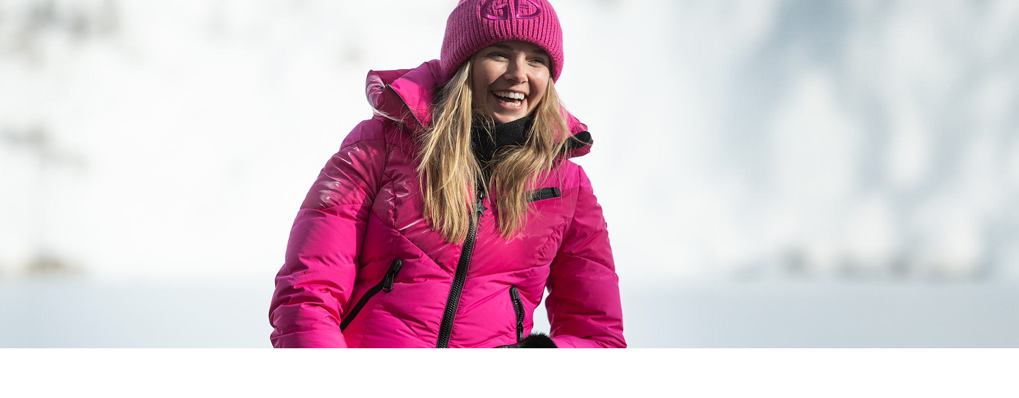 Shop gifts for her. A woman in a pink ski jacket and hat with a snowy backdrop 