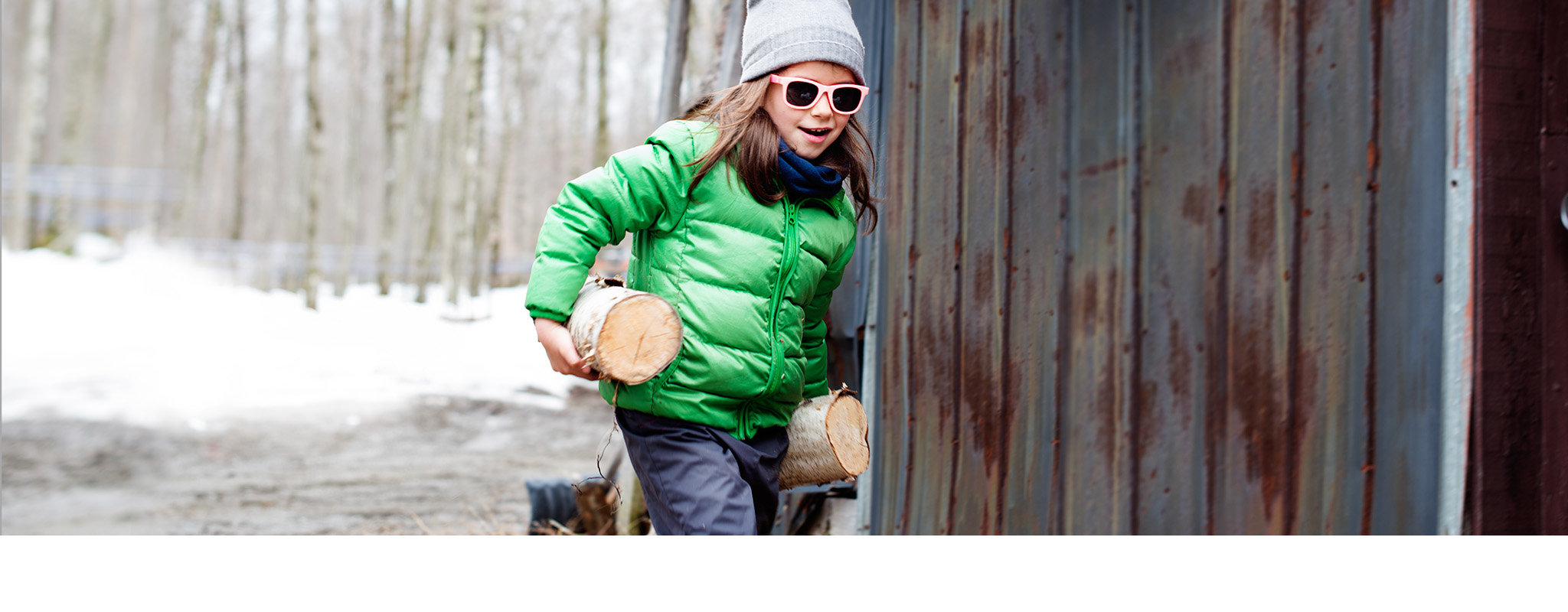 Shop gifts for kids. A child carrying logs wearing a winter jacket, beanie and sunglasses