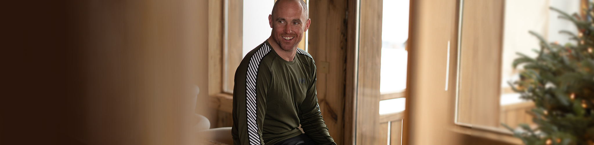 Shop Gifts under £100. A man next to a Christmas tree wearing a baselayer