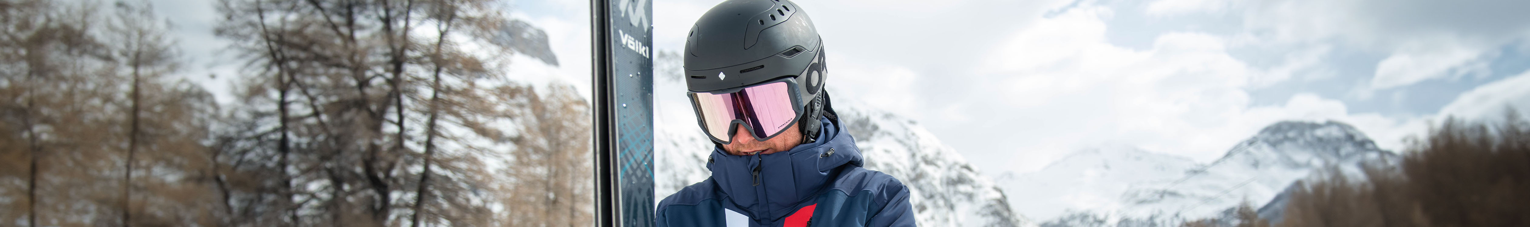 Shop helmets. A man is wearing a black helmet, googles and a jacket, holding skis.