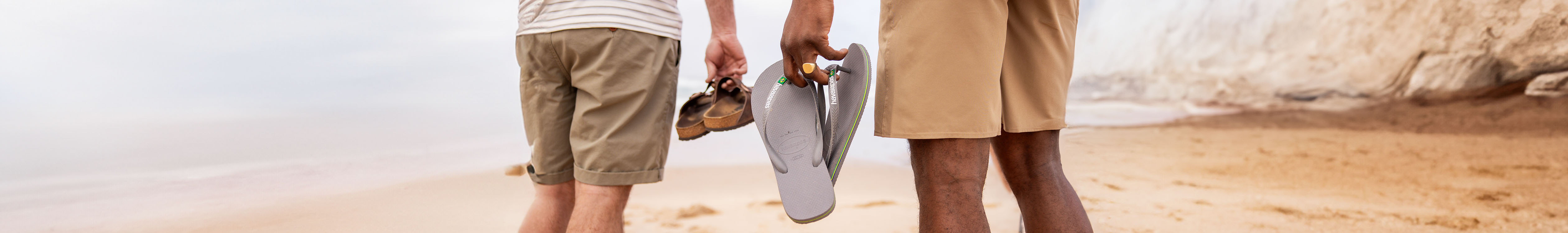 Sandals collection. Two people in shorts are holding their flip-flops and sandals, while walking barefoot on a sandy beach. 