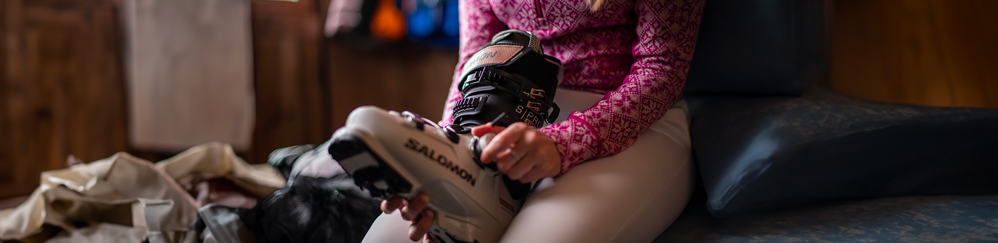 Shop snowsports footwear. A person is holding a ski boot to put it on, while sitting on a bench inside