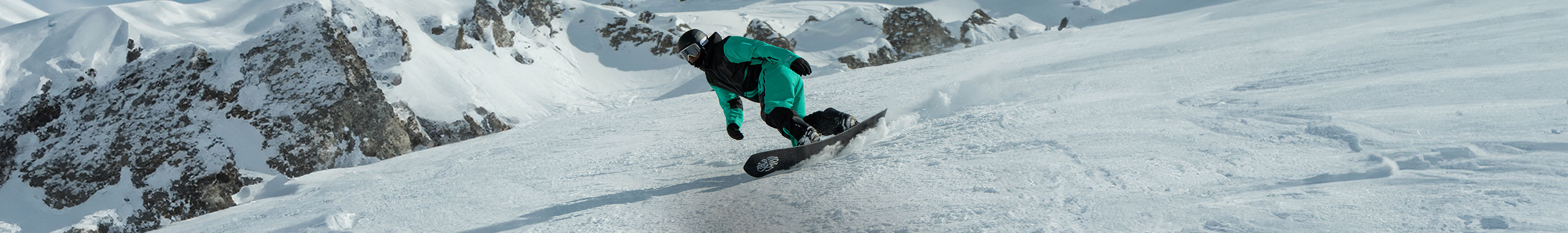 Shop Snowboards. A person in green snowboard jacket and pants snowboarding down the slope 