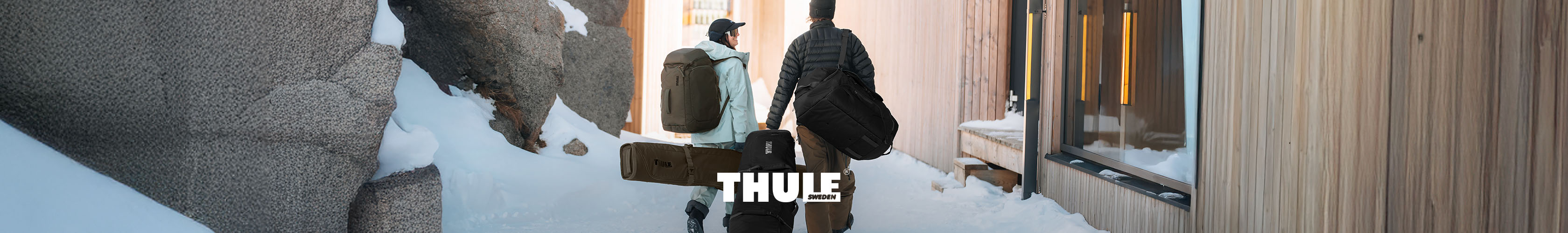 Shop Thule collection. Two people with backpacks and ski bags walking through snowy ski resort