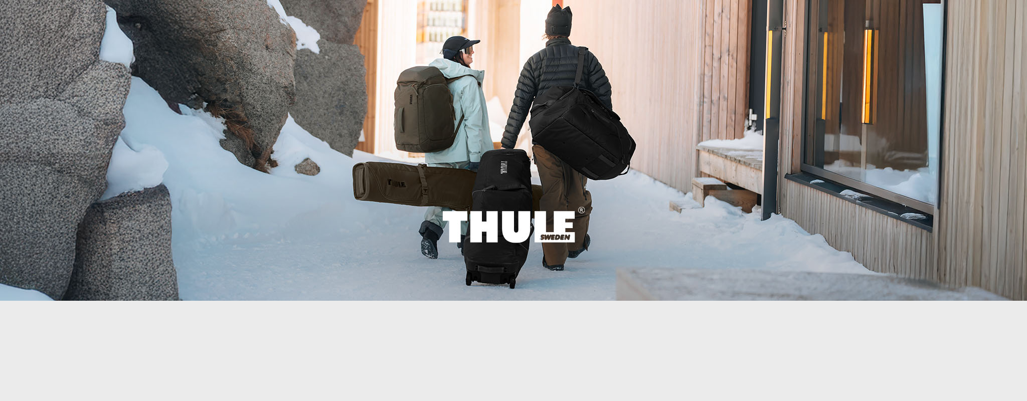 Shop Thule collection. Two people with backpacks and ski bags walking through snowy ski resort