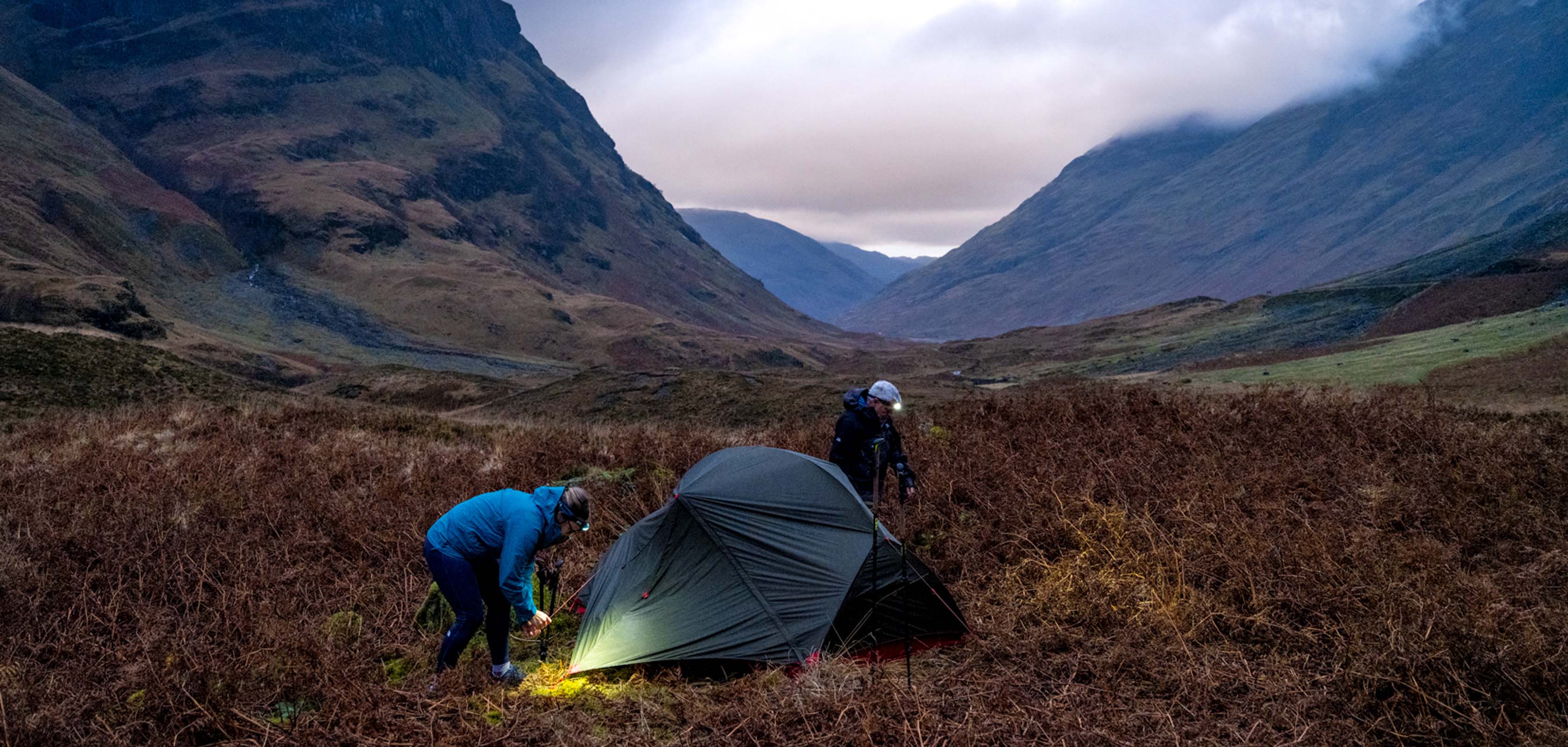 Two peopleare pitching a tent somewhere outdoors, wearing walking gear and head torches on their heads.