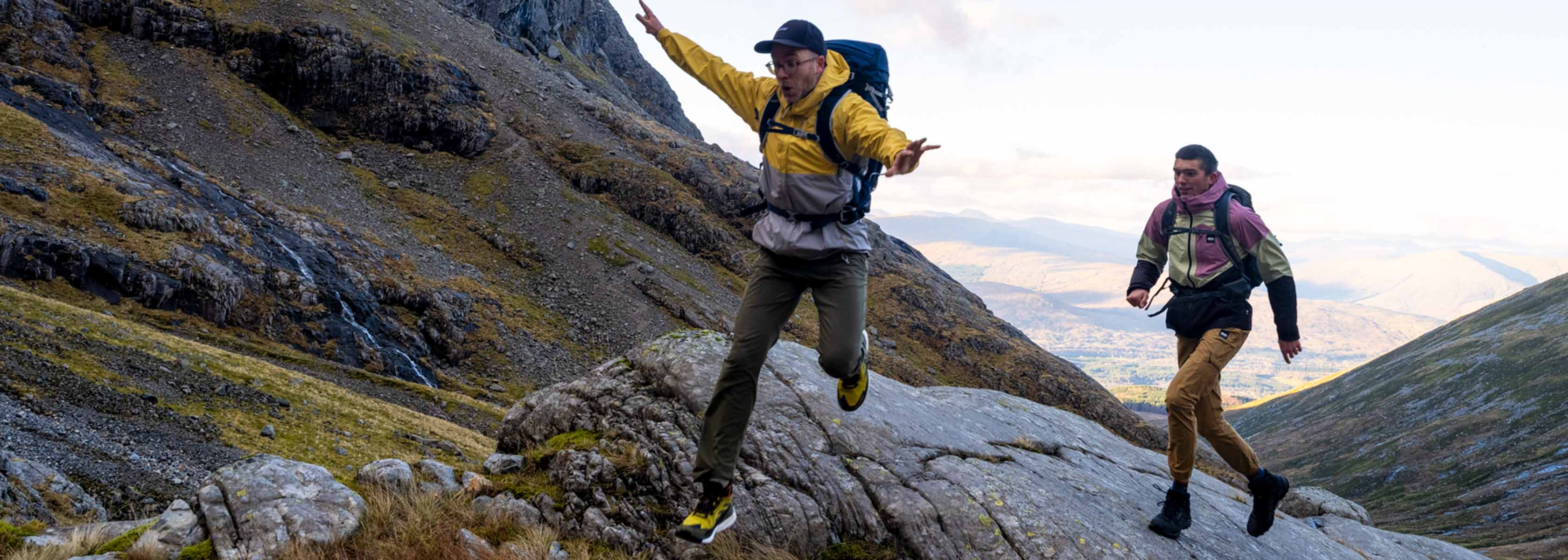 Two men are alking fast on big rocks, wearing walking gear and rucksacks.