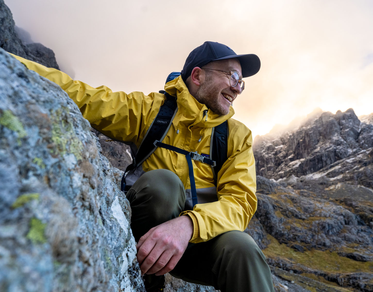 A man is smiling somewhere surraunded by the mountains, wearing a blueyellow waterproof jacket, a cap, and a backback.